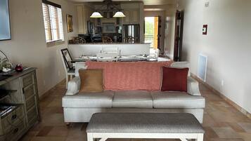 A spacious living room with a grey sofa, a white bench, and a kitchen in the background.