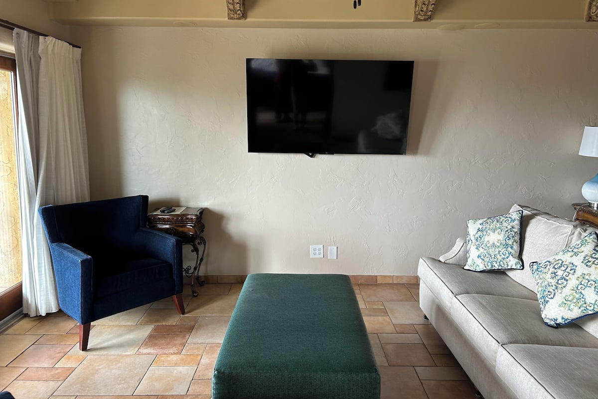 A living room with a flat-screen TV mounted on the wall, a blue armchair, a green ottoman, and a light-colored sofa with patterned cushions.