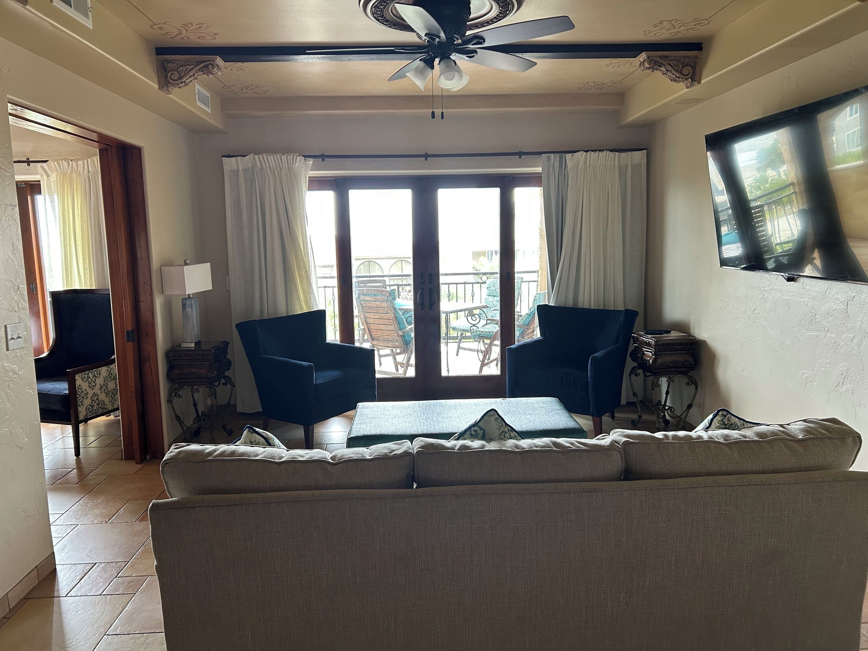 A living room with a sofa, two armchairs, a table, a ceiling fan, and a large window with curtains.