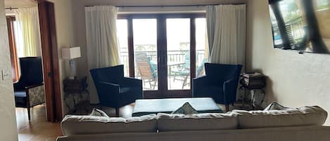 En stue med sofa, to lænestole, et bord, en loftventilator og et stort vindue med gardiner.