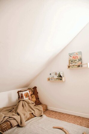 Un dormitorio abrigado en un ático, con techo inclinado, una cama con manta marrón y dos estantes empotrados con libros y una pequeña fotografía.