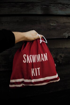 Eine Hand hält eine rote Tasche mit dem Aufdruck 'SAWMAN KIT' vor einem Holzhintergrund.