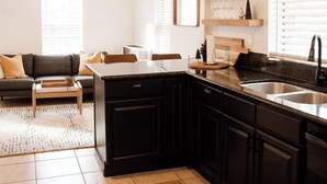 A modern kitchen with black cabinets, a granite countertop, and a built-in sink. There is a dining area with a sofa and a coffee table.
