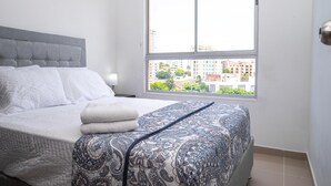 A neatly made bed with a patterned bedspread and pillows, a grey upholstered headboard, and a window with a view of buildings and greenery.