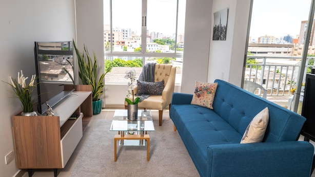 A modern living room with a blue sofa, a glass coffee table, a TV on a wooden stand, and a view of the city through large windows.
