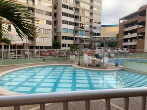 Ein Schwimmbecken mit Schachbrettfliesen, umgeben von einem Zaun, neben einem mehrstöckigen Gebäude mit Balkonen.