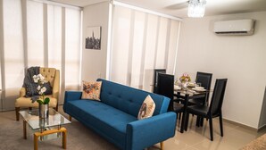 A living room with a blue sofa, a glass coffee table, a beige armchair, and a dining area with a table and chairs.