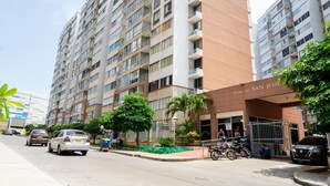 A multi-story hotel with balconies, a gated entrance, and a parking area with cars and motorcycles.