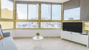 A modern living room with a large window, a flat-screen TV, a small round table with a plant, and a grey sofa. - Home2Book Design Las Canteras2 Beach (Las Palmas de Gran Canaria)