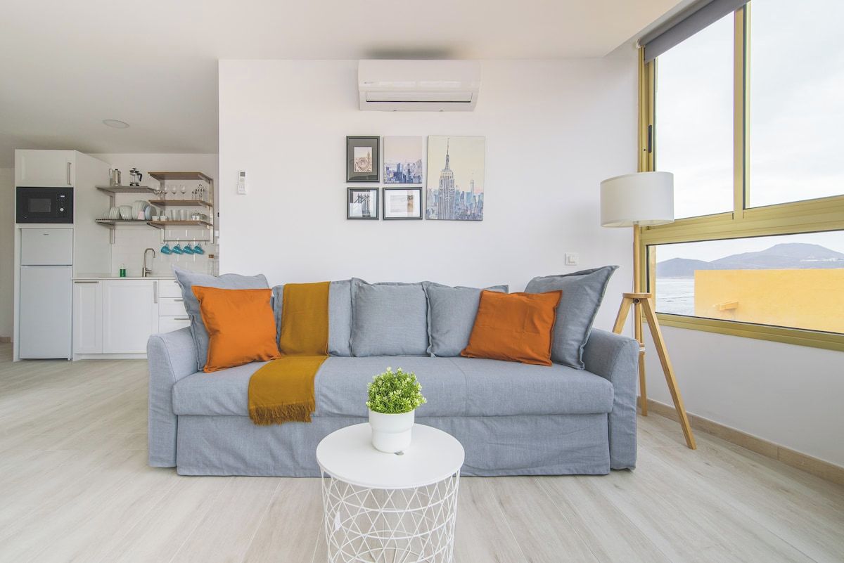 A living room with a grey sofa, a small round table with a plant, a white air conditioning unit, and a window with a view.