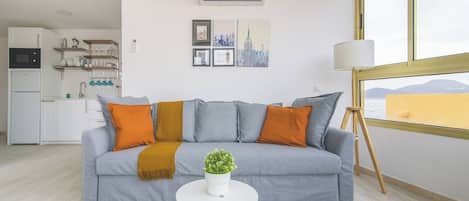 A living room with a grey sofa, a small round table with a plant, a white air conditioning unit, and a window with a view.