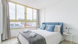 A hotel room with a large window, a bed with a grey and white duvet, blue pillows, and a white bedside table with a lamp. - Home2Book Design Las Canteras2 Beach (Las Palmas de Gran Canaria)