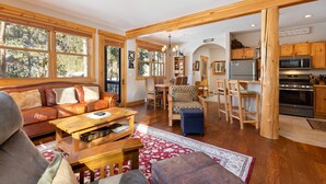 A spacious living room with a leather sofa, wooden coffee table, and a red patterned rug. There is a dining area with wooden chairs and a table, and a kitchen with wooden cabinets and a microwave.