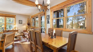 A dining area with a wooden table and chairs, large windows, and a chandelier.