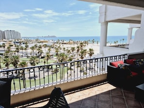 A balcony with a view of a beach, palm trees, and a distant cityscape.