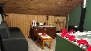 Une pièce mansardée chaleureuse avec un plafond en bois, un mur vert, une cheminée, un canapé, une table ornée d’un vase de fleurs rouges et un sol carrelé.