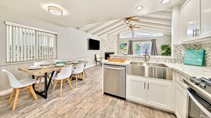 A modern kitchen with white cabinetry, a wooden dining table with chairs, a built-in dishwasher, and a ceiling fan. - Perfect 3 Bedroom Home between the Bay and Beach (Newport Beach)
