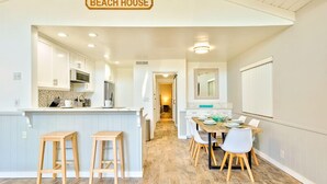 A kitchen with white cabinets, a wooden island with stools, and a dining area with a table and chairs. - 6 Bedroom Home with Rooftop Deck Between the Bay & the Beach (Newport Beach)