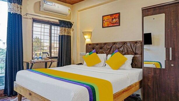 A hotel room with a bed, a bedside table, a lamp, a window with curtains, an air conditioning unit, and a painting on the wall. - Itsy Hotels Punest (Pune)