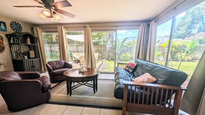 A sunlit living room with a ceiling fan, leather sofas, a coffee table, and a view of a backyard through large windows. - Artist owned home near beaches (Venice)