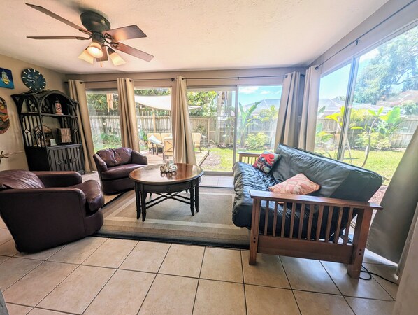 A sunlit living room with a ceiling fan, leather sofas, a coffee table, and a view of a backyard through large windows.