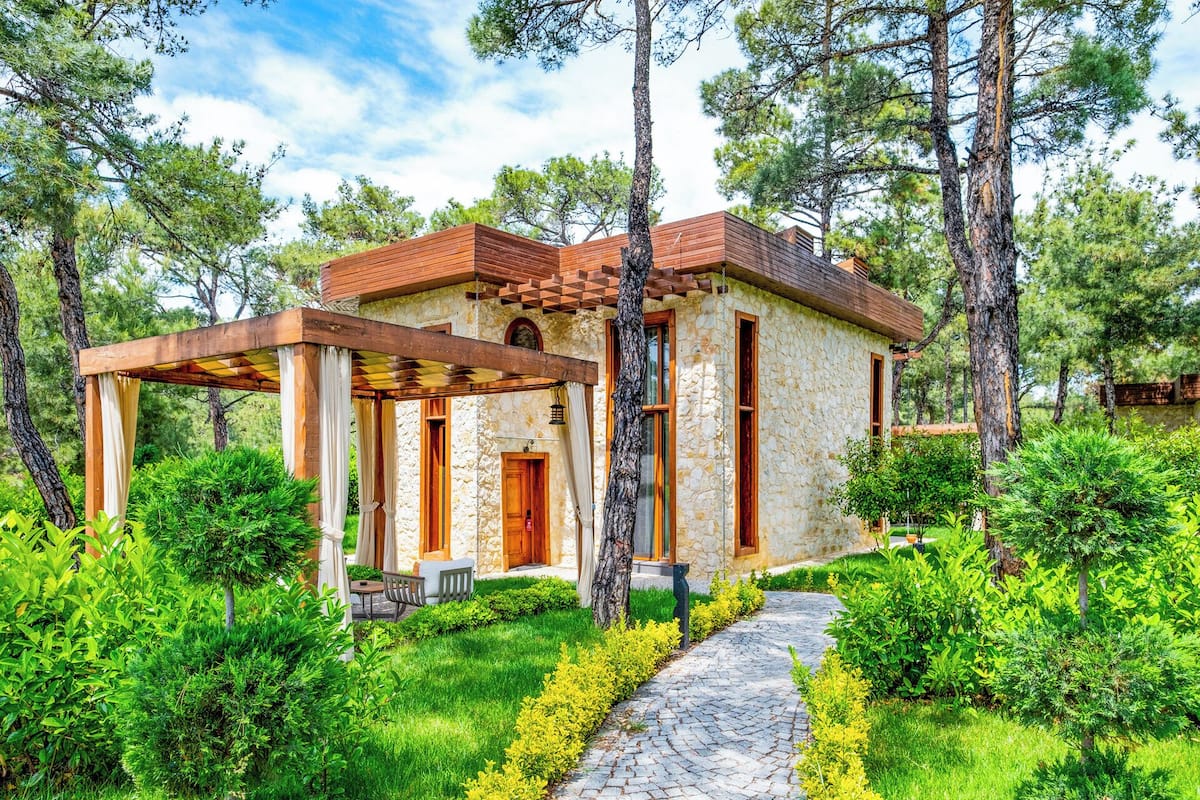 Ein Steinhaus mit einer Holzpergola, umgeben von üppigem Grün und Bäumen.