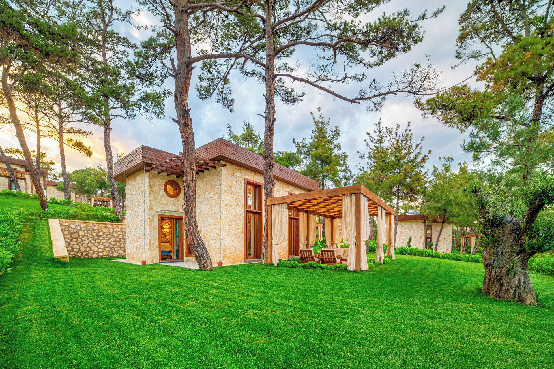 A stone building with a wooden pergola, surrounded by lush green grass and trees.