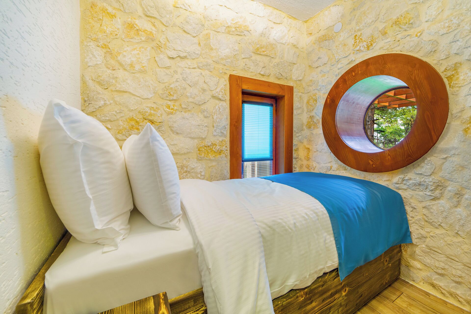 Un lit avec une literie blanche et un élément décoratif bleu, un mur en pierre, une porte en bois et une fenêtre ronde donnant sur un paysage verdoyant.