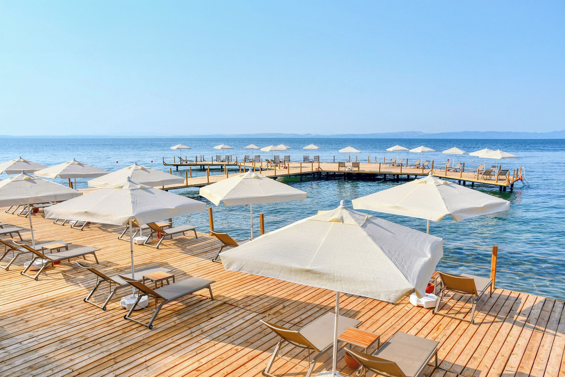 Una terraza de madera con tumbonas y sombrillas, un muelle que se adentra en el mar, y cielos despejados.
