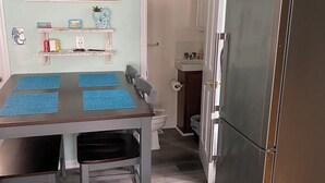 A kitchen with a stainless steel refrigerator, a dining table with blue placemats, and a painting of palm trees on the wall. - Cozy Cottage 3 blocks to Town 7 to Beach 4 Blocks to Asbury (ocean grove)