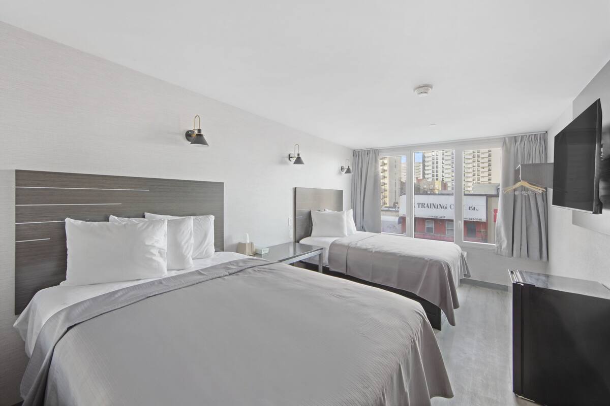 A hotel room with two beds, a desk, a TV, and a window with a view of buildings.