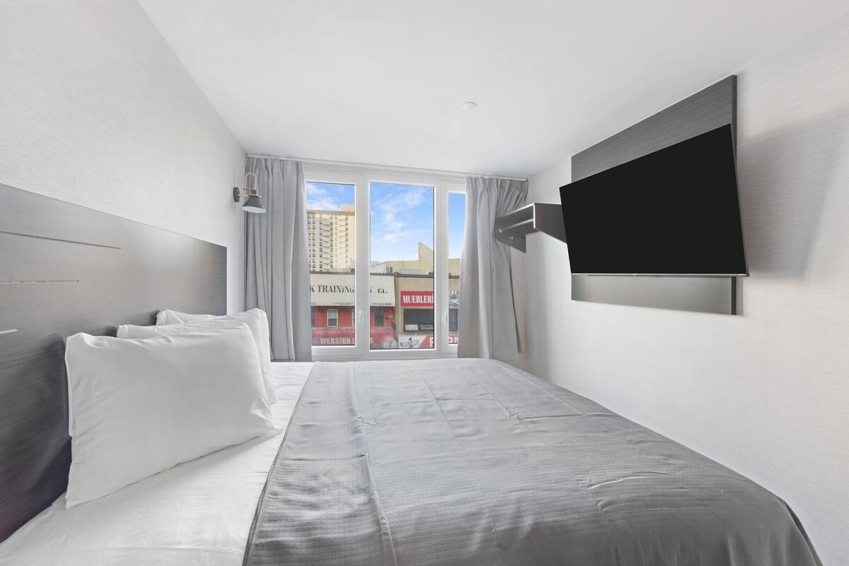 A modern hotel room with a large bed, a flat-screen TV mounted on the wall, and a view of the city through large windows.