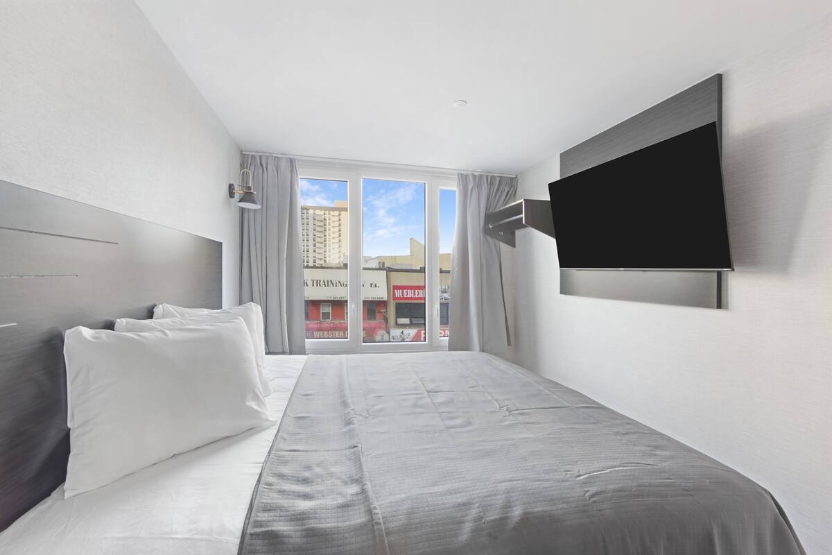 A modern hotel room with a large bed, a flat-screen TV mounted on the wall, and a view of the city through large windows.