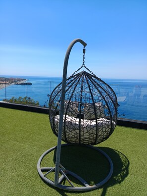 A hanging chair with a metal frame and netting, placed on artificial grass with a view of the sea and buildings in the background.