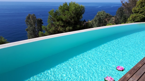 A clear blue infinity pool with wooden deck and pink lotus floats, overlooking a sea view.