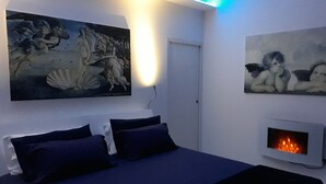 A bedroom with a bed, two wall-mounted paintings, a wall-mounted fireplace, and a ceiling light.