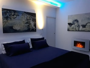Quarto com uma cama, duas pinturas penduradas na parede, lareira na parede e uma luminária de teto.