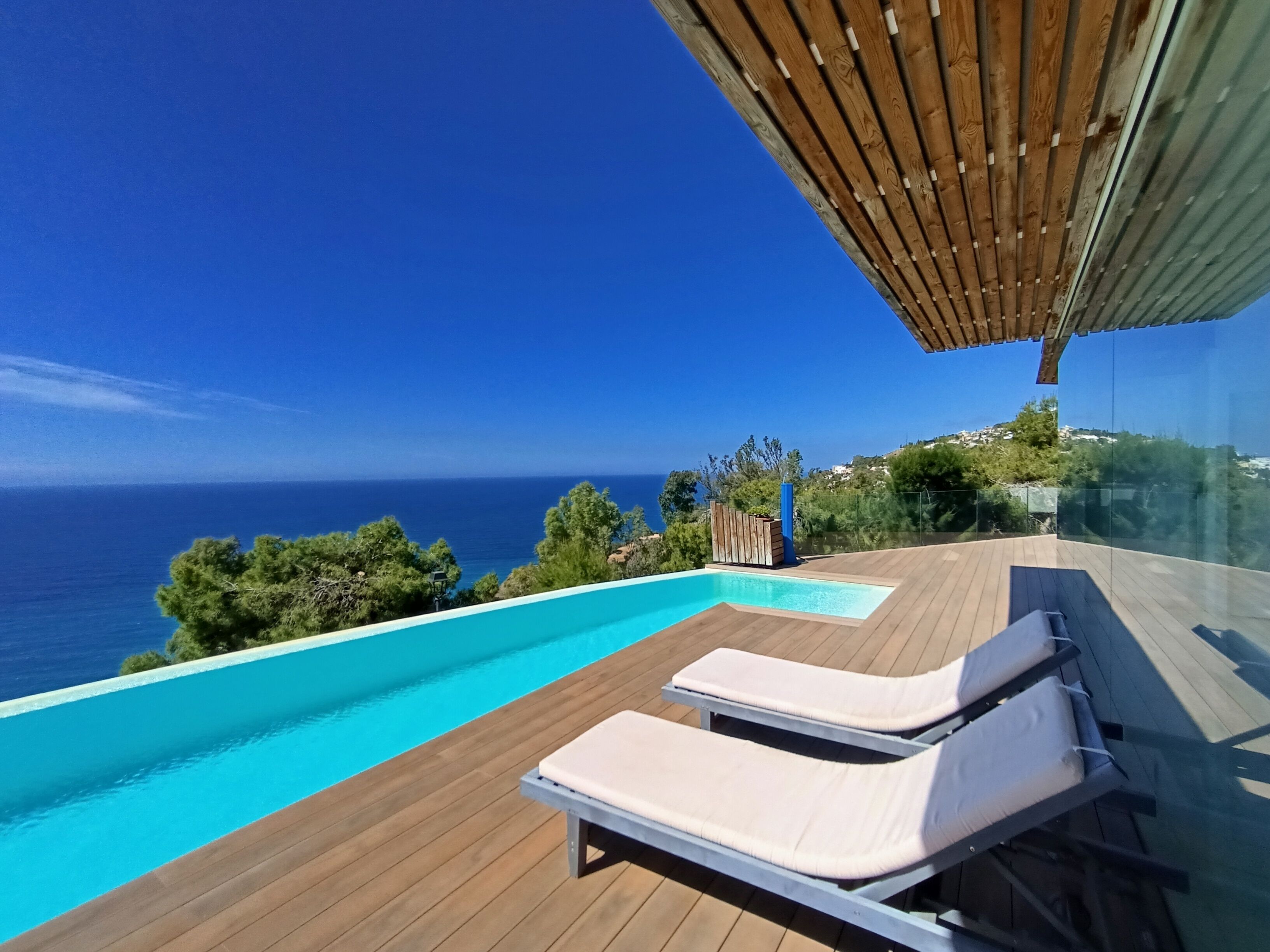A modern outdoor swimming pool with wooden deck and lounge chairs, overlooking a coastal landscape.