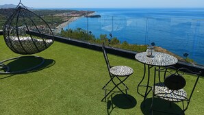 A terrace with artificial grass, a hanging chair, and two tables with mosaic tops overlooking a coastal view.