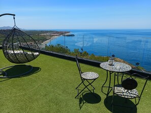 Une terrasse avec un revêtement de sol en gazon synthétique, un fauteuil suspendu et deux tables dont les plateaux sont décorés de mosaïques, le tout offrant une vue sur la côte.