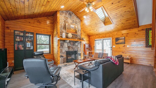 L’intérieur chaleureux d’un chalet avec une cheminée en pierre, des murs en bois, un ventilateur de plafond et un coin salon comprenant un canapé et des fauteuils.