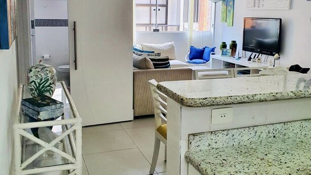A modern kitchen with a granite countertop, a dining area with a white table and chairs, and a living room with a sofa and a TV.