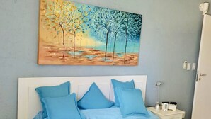 A bedroom with a bed, blue bedding, a painting of trees, and an air conditioning unit.