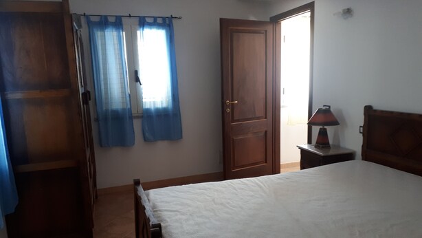 A bedroom with a bed, a wooden wardrobe, a nightstand with a lamp, and a door with curtains. - Pink House (Sardegna)
