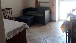 A cozy living area with a sofa, a small table, and a chair. There is a window with curtains, a door, and a tiled floor. - Pink House (Sardegna)
