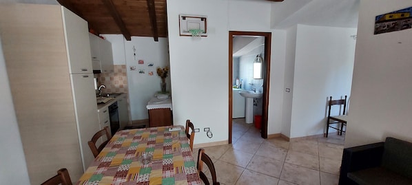 A dining area with a table covered in a patchwork tablecloth, wooden chairs, and a kitchen in the background.