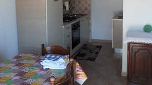 A compact kitchen with a refrigerator, stove, oven, and sink. There is a dining table with a quilted tablecloth and a chair. The room has wooden beams on the ceiling and a small cabinet with a green object on top. - Pink House (Sardegna)