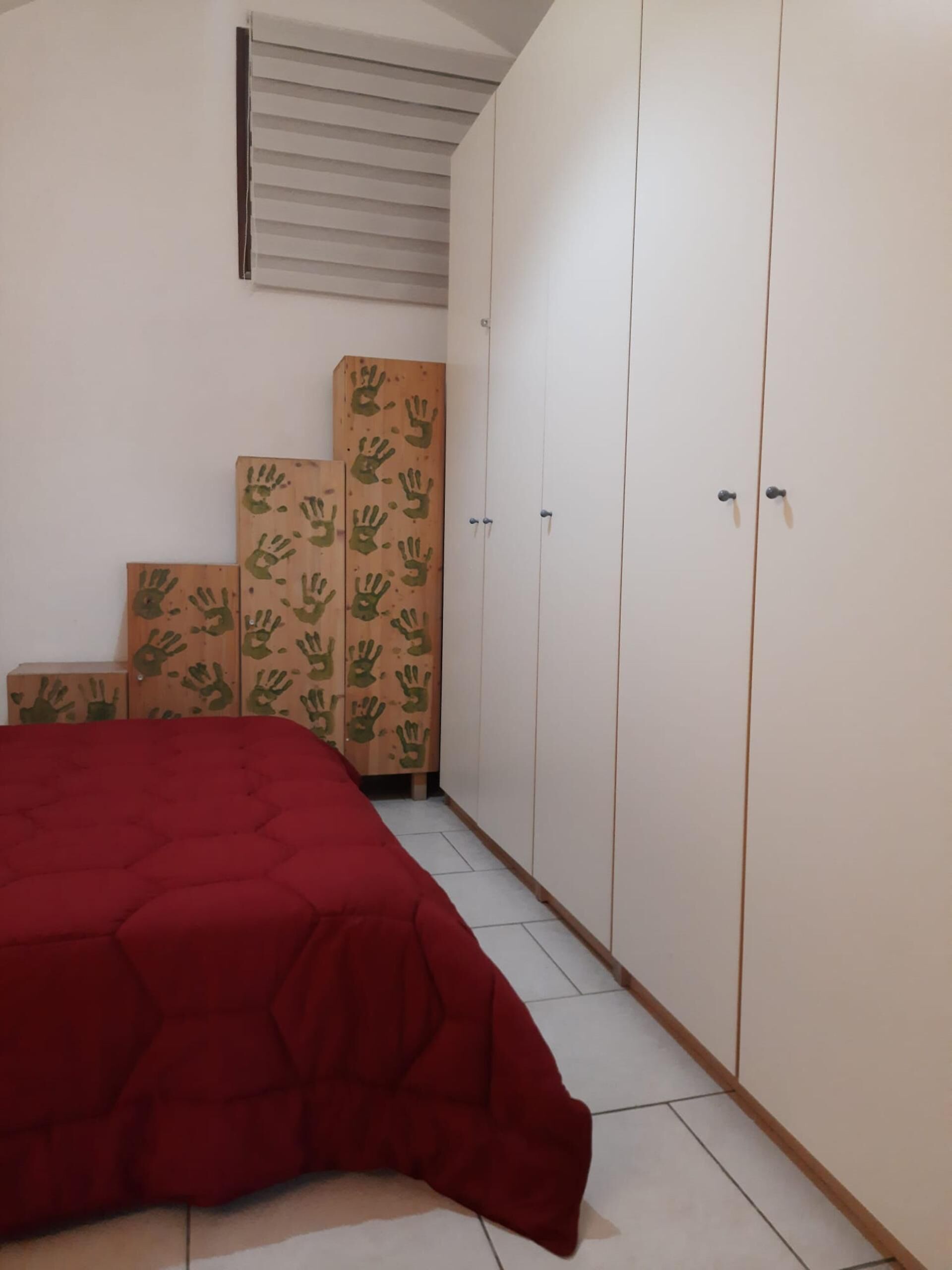 Une chambre avec une couvre-lit rouge, une armoire blanche et des cartons ornés d’empreintes de mains.