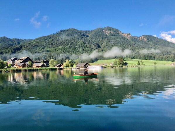 Un lago tranquilo con una hilera de casas de madera tradicionales a lo largo de la orilla, un bote con una persona remando y una cadena montañosa al fondo.