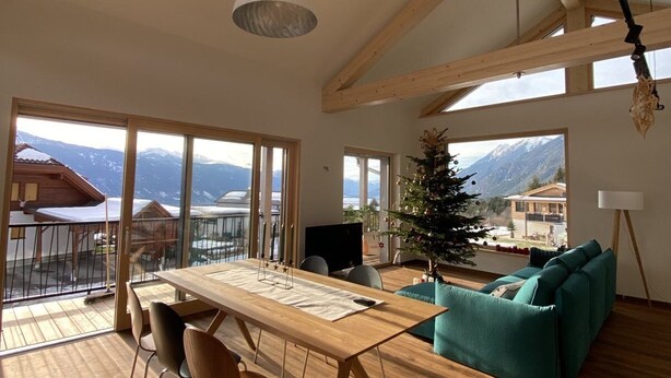 A spacious living area with a wooden dining table, chairs, and a sofa. Large windows offer views of mountains and a balcony. - Eco-Chalet Matschiedl
Luxury mountainside Eco Chalet with stunning view -  (Kärnten)